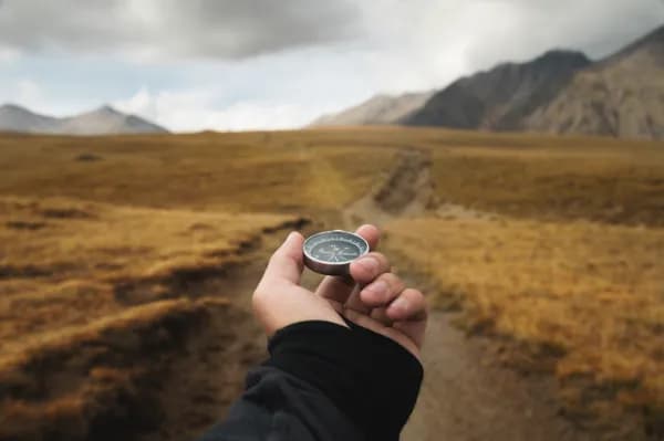 Hand holding a compass toward a path through an open landscape.