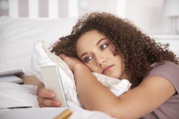 Young woman resting on a bed while holding a smartphone.